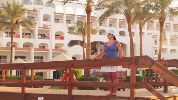 Brunetka Židovka v modrém klobouku stojí na mostě a obdivuje palmy na pozadí hotelu. — Stock video