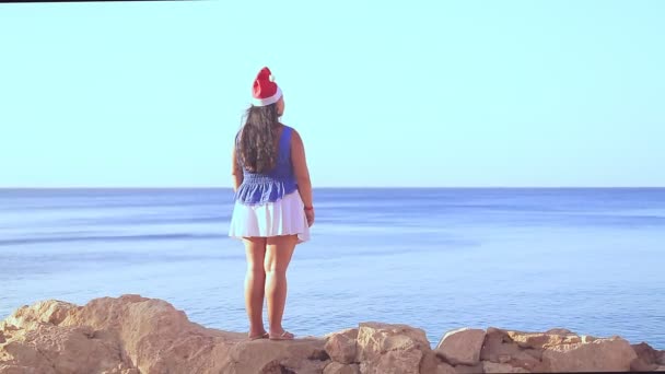 Una mujer morena en un sombrero de Santa camina por la costa rocosa admirando la superficie del mar — Vídeos de Stock
