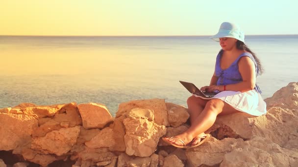 Una mujer morena con un sombrero azul en una costa rocosa al sol trabaja remotamente con un portátil — Vídeo de stock