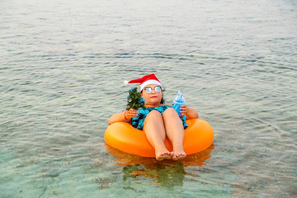 A woman in a swimming ring in a Santa Claus hat with a cocktail and a small Christmas tree in her hand swims in the sea on holidays. — Stock Photo, Image