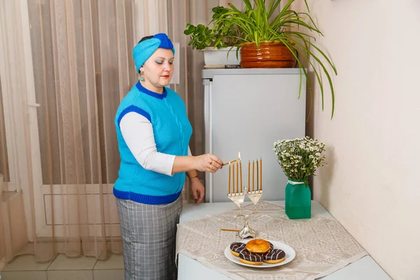 Еврейка в синем жилете и тюрбан в столовой зажигают свечи на Хануке на столе рядом с пончиками и вазой с цветами. — стоковое фото