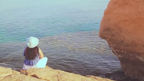 Brunetka v modrém klobouku sedí zády na kamenech a obdivuje moře. — Stock video