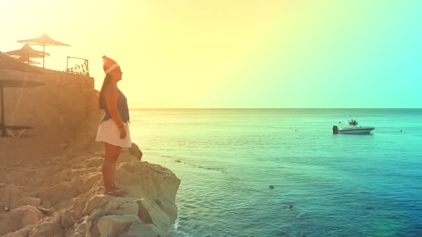 Una mujer morena con un sombrero de Santa Claus está en la orilla del mar en una roca admirando la llamarada del sol — Vídeos de Stock