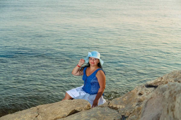 Beyaz etekli ve deniz kıyısında güneş şapkalı genç bir kadın doğaya hayran bir taşın üzerinde oturuyor. Yatay fotoğraf