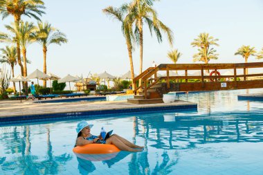 Yüzme havuzunda güneş şapkalı esmer bir kadın kokteyl ve dizüstü bilgisayarla palmiye ağaçlarının arka planında görüntülü iletişim kuruyor. Yatay fotoğraf
