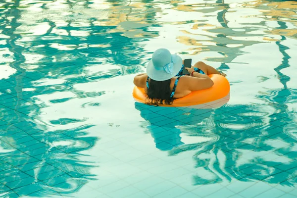 Yüzme havuzunda mayo ve güneş şapkası takmış akıllı telefonlu bir kadının arka plan görüntüsü. Yatay fotoğraf