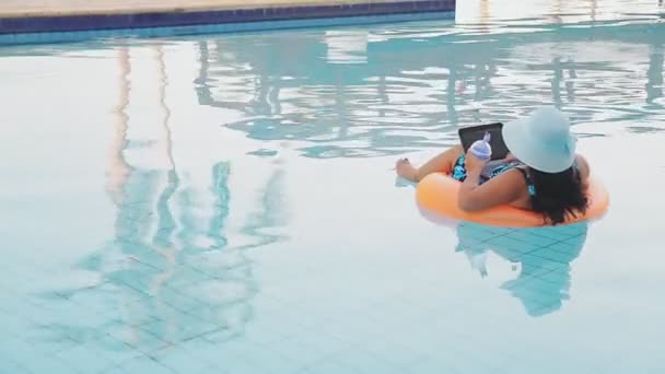 Mulher com um chapéu de sol e um maiô na piscina em um círculo de natação com um smartphone bebe um coquetel com as costas para a câmera — Vídeo de Stock