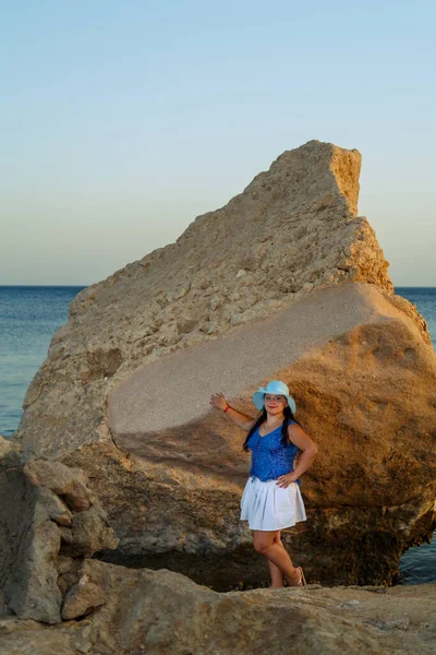 Μια νεαρή γυναίκα σε μια λευκή φούστα και ένα καπέλο ήλιο στέκεται στην ακτή μεταξύ των βράχων με φόντο τη θάλασσα στο ηλιοβασίλεμα. — Φωτογραφία Αρχείου