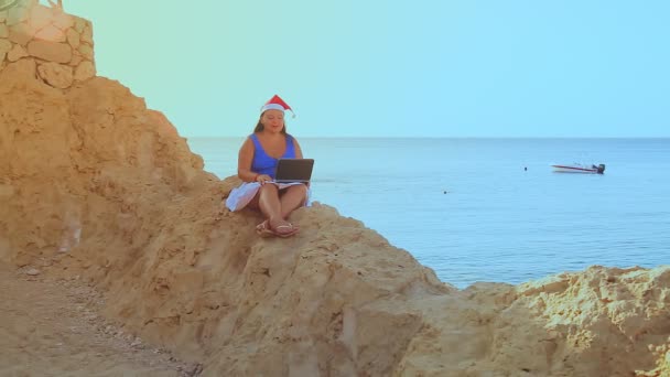 Una donna bruna in un cappello di Babbo Natale si siede sulla riva del mare su una roccia lavora con un computer portatile — Video Stock