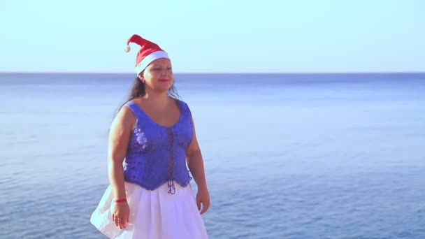 Een brunette vrouw in een kerstman hoed loopt op de rotsen door de zee en bewondert de natuur — Stockvideo