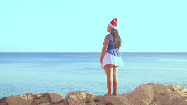 Uma mulher morena em um chapéu de Papai Noel caminha pela costa rochosa admirando a superfície do mar — Vídeo de Stock