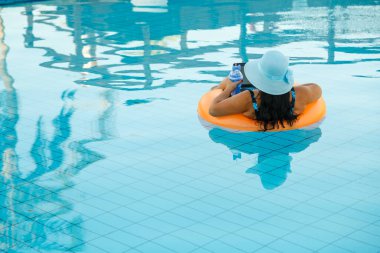 Yüzme havuzunda güneş şapkalı bir kadın elinde dizüstü bilgisayarla arkadan ateş ederken gevşiyor..