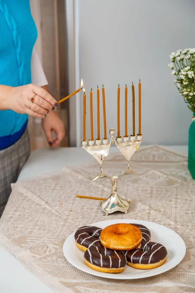 Рука еврейки в столовой на кухне зажигает свечи на Хануке на столе рядом с пончиками и вазой с цветами. — стоковое фото