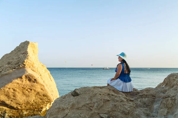 Μια νεαρή μελαχρινή γυναίκα με λευκή φούστα και μπλε καπέλο στην παραλία κάθεται με την πλάτη της στην κάμερα.. — Φωτογραφία Αρχείου