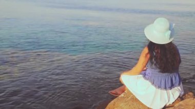 Mavi şapkalı esmer bir kadın sırtı kayaların arasında oturur ve denize ve günbatımına hayran kalır..