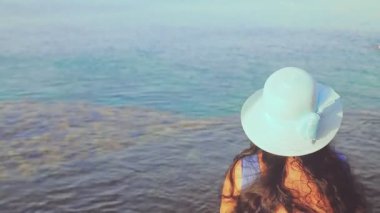Mavi şapkalı esmer bir kadın kameraya arkası dönük denize hayran.