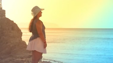 Kayalık bir sahilde mavi şapkalı esmer bir kadın, gün batımından alevler içinde, denize ve günbatımına hayran..