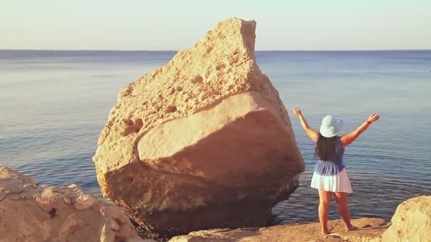 A brunette woman in a blue hat among the rocks by the sea admires nature with her arms outstretched — Video Stock
