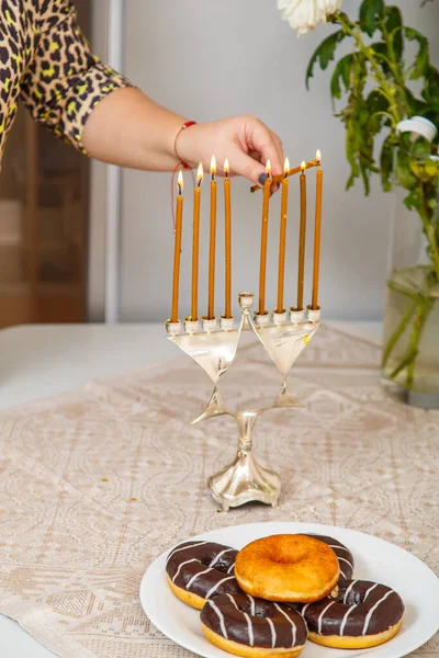 Рука еврейки зажигает свечи на Хануке на столе рядом с пончиками в тарелке и вазой с цветами. — стоковое фото