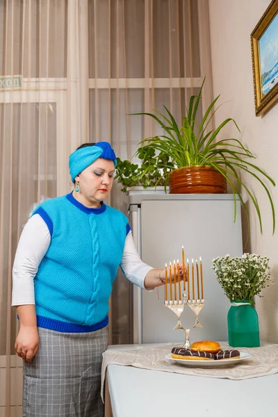 在餐厅厨房里，一位头戴头巾的犹太妇女在餐桌上点燃着蜡烛，旁边放着甜甜圈和一个花瓶. — 图库照片