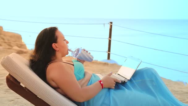 Une femme est allongée dans une chaise longue sur le bord de la mer à boire un cocktail et bavarde dans un gadget du côté — Video