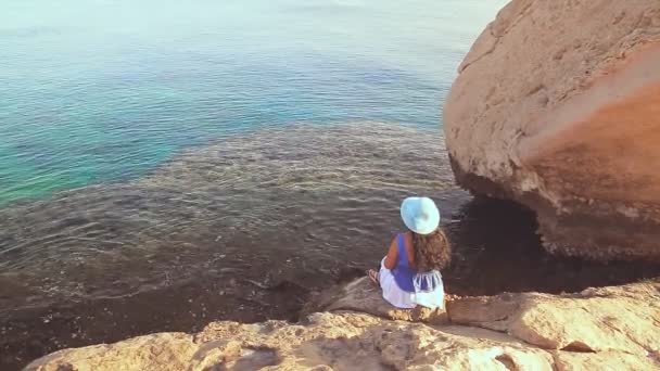 Ruskeaverikkö nainen sinisessä hatussa kivien keskellä ihailee merta ja auringonlaskua — kuvapankkivideo