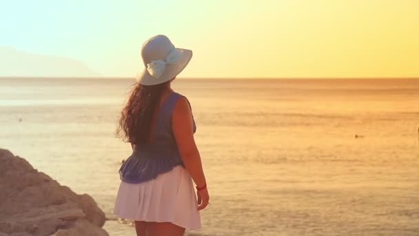 Una mujer morena en un sombrero azul en una costa rocosa, destello de la puesta del sol, admira el mar y la puesta del sol — Vídeos de Stock