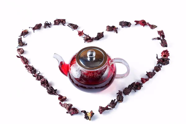 A teapot of hibiscus tea and hibiscus petals laid out in the shape of a heart on a white background.