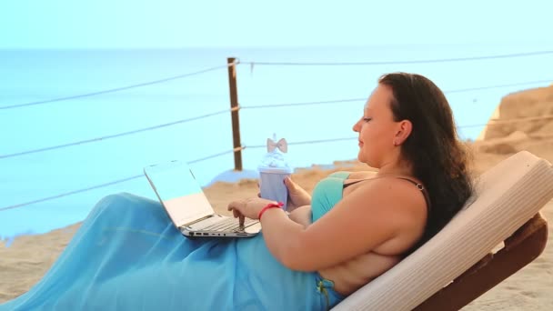 O femeie se află într-un șezlong de soare pe malul mării, bea un cocktail și vorbește într-un gadget din lateral. — Videoclip de stoc