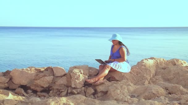 A brunette woman in a blue hat on a rocky seashore in the rays of the sun flare remotely works with a laptop. — Stock Video