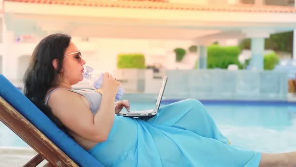 Een vrouw in een badpak en een pareo op een ligstoel bij het zwembad drinkt een cocktail en werkt op afstand. — Stockvideo