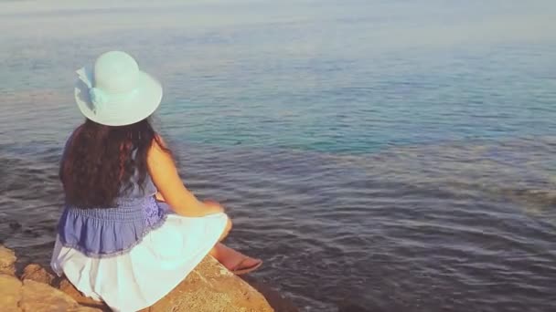 A brunette woman in a blue hat sits with her back among the rocks admiring the sea and the sunset — 图库视频影像