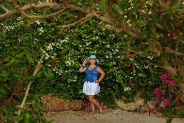 Beyaz etek ve güneş şapkalı esmer bir kadın çiçek açan beyaz begonvilin yanında duruyor..