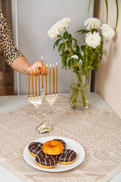 The hand of a Jewish woman lights candles in Hanukkah on the table next to festive donuts in a plate and a vase of flowers. — 图库照片