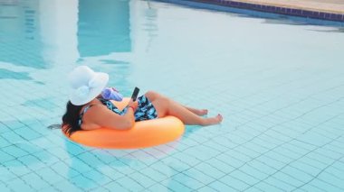 joyful woman in a sun hat and a swimsuit in the pool in a swimming circle with a smartphone drinks a cocktail and rests