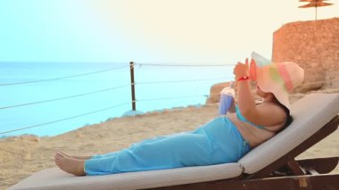 Pareo giymiş bir kadın ve deniz kıyısındaki bir şezlongda şapka takan bir kadın denize bir kokteyl içiyor.