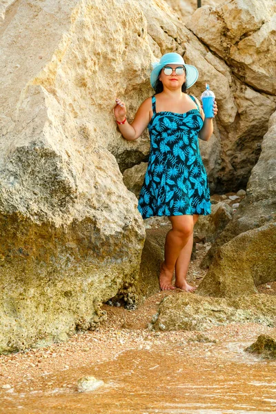 Mulher judia feliz em um vestido azul e chapéu de palha e óculos nas rochas com um coquetel na mão. — Fotografia de Stock