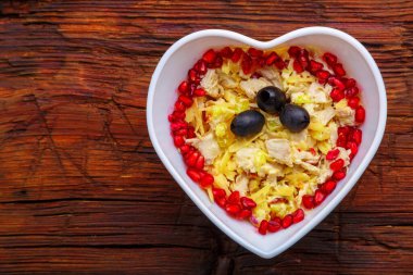 Tavuk göğsü ve ananasla şişirilmiş salata, nar ve zeytinlerle süslenmiş ahşap bir masada kalp salata kasesi..