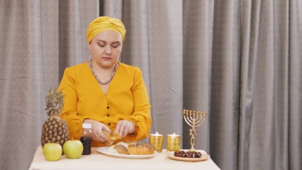Еврейская женщина в традиционном головном уборе ест халу после благословения во время субботнего собрания — стоковое видео
