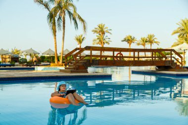 Havuzda güneş şapkası takmış, yüzme havuzunda kokteyl ve palmiye ağaçlarının arka planında dizüstü bilgisayarı olan Yahudi bir kadın. Yatay fotoğraf