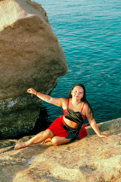 En brunett kvinna i en röd orientalisk dans kostym sitter på en klippa mot bakgrund av havet. — Stockfoto