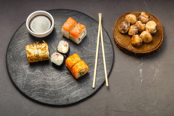 Rolls with fish and shrimp on a round board next to sticks, soy sauce and baked champignons on a dark background. — Stock Photo, Image