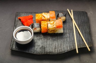 Çubukların, soya sosunun ve arka planda zencefilin yanında siyah bir tahtanın üzerinde çeşitli rulo ekmekler. Yatay fotoğraf