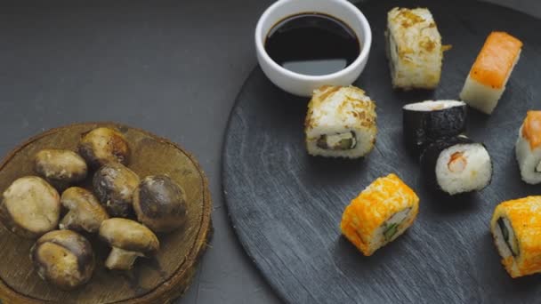 Rouleaux assortis sur une planche ronde à côté de la sauce soja et champignons cuits au four sur un fond noir — Video