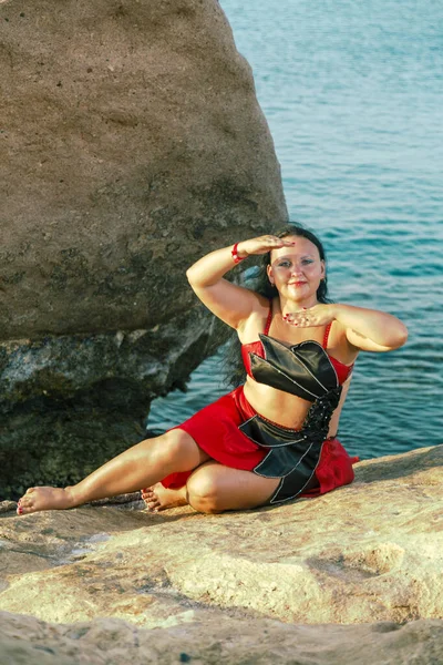 Una morena vestida con un traje de baile oriental rojo se sienta sobre una roca en el fondo del mar. — Foto de Stock