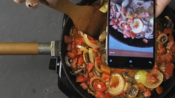 A womans hand mixes vegetables fried in a frying pan with a spatula and shoots a blog on the phone — Stock Video