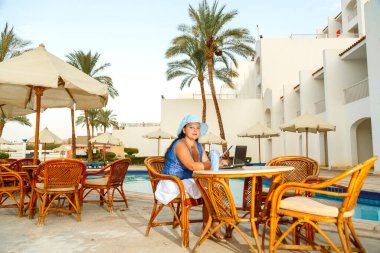 Otelin önündeki bilardo masasında şapkalı genç bir kadın kokteyl içer ve uzaktan bilgisayarla çalışır..