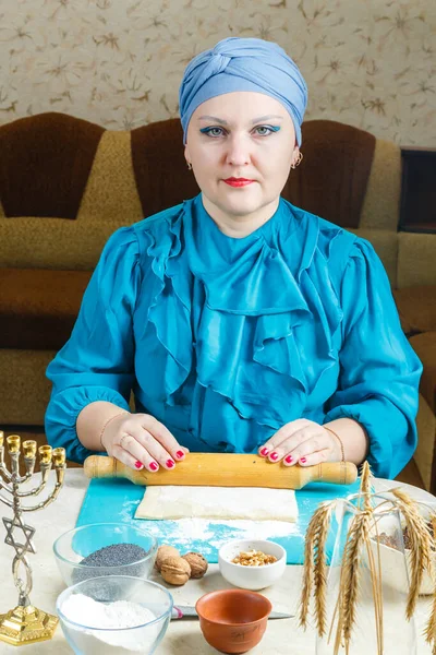 一位身穿传统头饰的犹太妇女在餐桌上用滚动式别针取出面团，用罂粟籽和李子做"哥根塔什饼干"，以庆祝普利姆节庆. — 图库照片