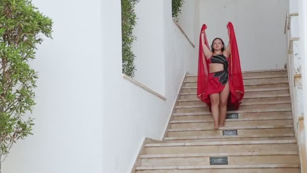 Uma mulher morena em um traje de dança oriental vermelho com um xale desce as escadas dançando — Vídeo de Stock