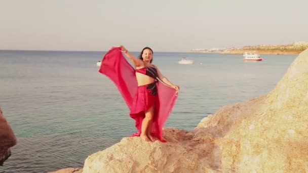 Una morena vestida con un traje de baile oriental rojo baila con un chal sobre una roca junto al mar. — Vídeo de stock